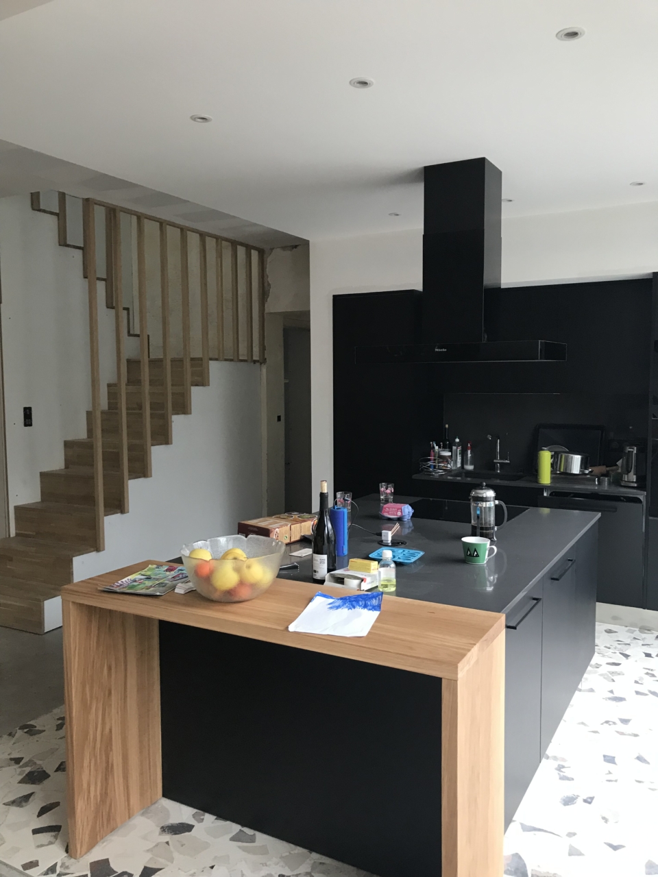Cuisine rénovée sur mesure à Bordeaux avec îlot central noir, coin repas en bois et escalier sur mesure en bois pour accéder à la surélévation de la maison